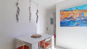 Una habitación con una mesa blanca, dos sillas, un cuadro de un pueblo costero y peces de juguete colgando.