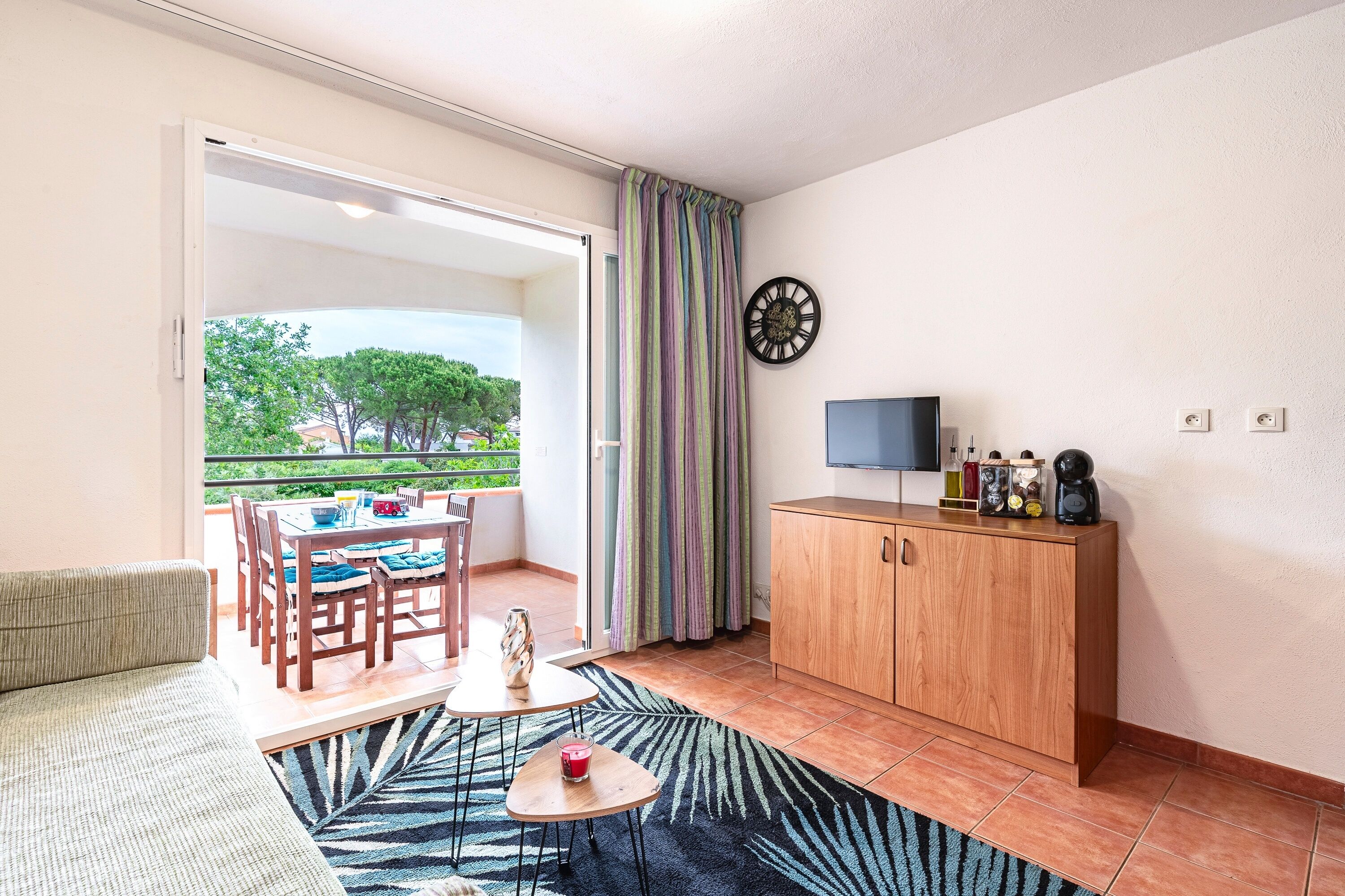 Ein Zimmer mit einer Couch, einem Esstisch mit Stühlen, einem kleinen Couchtisch, einem Schrank, einem Fernseher und Blick ins Grüne.