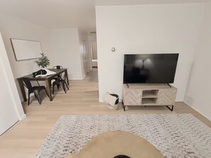 Ein Wohnzimmer mit Fernseher auf einem Holzständer, einem Esstisch mit Stühlen, einer Topfpflanze und einem gemusterten Teppich.