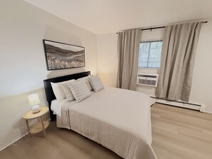 Ein Schlafzimmer mit einem Bett, einem Nachttisch mit Lampe, einem Fenster mit Vorhängen, einer Klimaanlage und einem gerahmten Bild an der Wand.