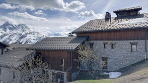 Ein traditionelles Chalet mit Steinwänden und Holzbalken, vor einer Kulisse aus schneebedeckten Bergen und einem teils bewölkten Himmel.