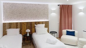 Ein Hotelzimmer mit zwei Betten, einem kleinen Tisch mit Lampe, einem Sessel und einem großen Fenster mit Vorhängen.