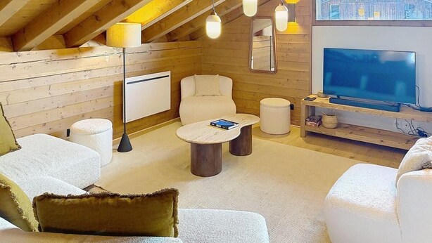 Ein gemütliches Zimmer mit Holzbalkendecke, einem Flachbildfernseher, einem weißen Sofa, einem runden Couchtisch und einem großen Fenster mit Blick auf ein Gebäude.