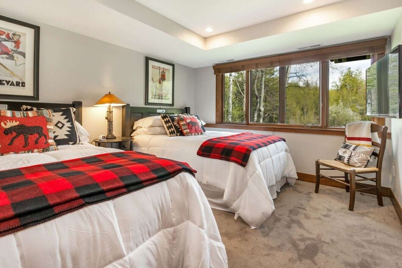 A hotel room with two beds, a chair, a window with a view of trees, and a lamp.