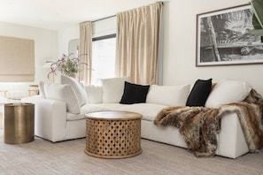 A modern living room with a white sofa, a round woven coffee table, and a large window with beige curtains. - The SouthOhana Retreat (Oceanside)