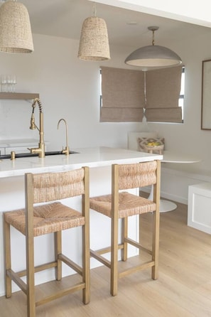 A modern kitchen with a white countertop, two wooden bar stools, a gold faucet, and two woven pendant lights. - The SouthOhana Retreat (Oceanside)