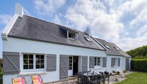 Ein zweistöckiges Haus mit geneigtem Dach, zahlreichen Fenstern mit Fensterläden, einem Essbereich mit Außenbestuhlung und einem kleinen Garten.