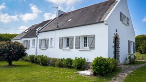 Ett vitt hus med gråa fönsterluckor, en stentrappa och en trädgård med buskar och ett träd.