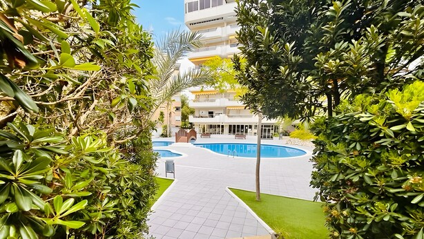 Un camino conduce a una piscina rodeada de abundante vegetación y un edificio de varios pisos al fondo.