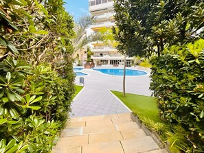 A pathway leading to a swimming pool surrounded by lush greenery and a multi-story building in the background. - SunAndBed Arquus II – Cozy Apartment with Pool in City Center, Steps from the Beach (Salou)