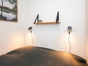 Un dormitorio con una cama, dos lámparas de pared, un estante con una planta pequeña y un cuadro enmarcado de un paisaje.