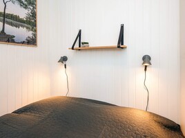 Un dormitorio con una cama, dos lámparas de pared, un estante con una planta pequeña y un cuadro enmarcado de un paisaje.