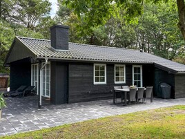 A black wooden cabin with a tiled roof, a chimney, and a patio area with a table and chairs.
