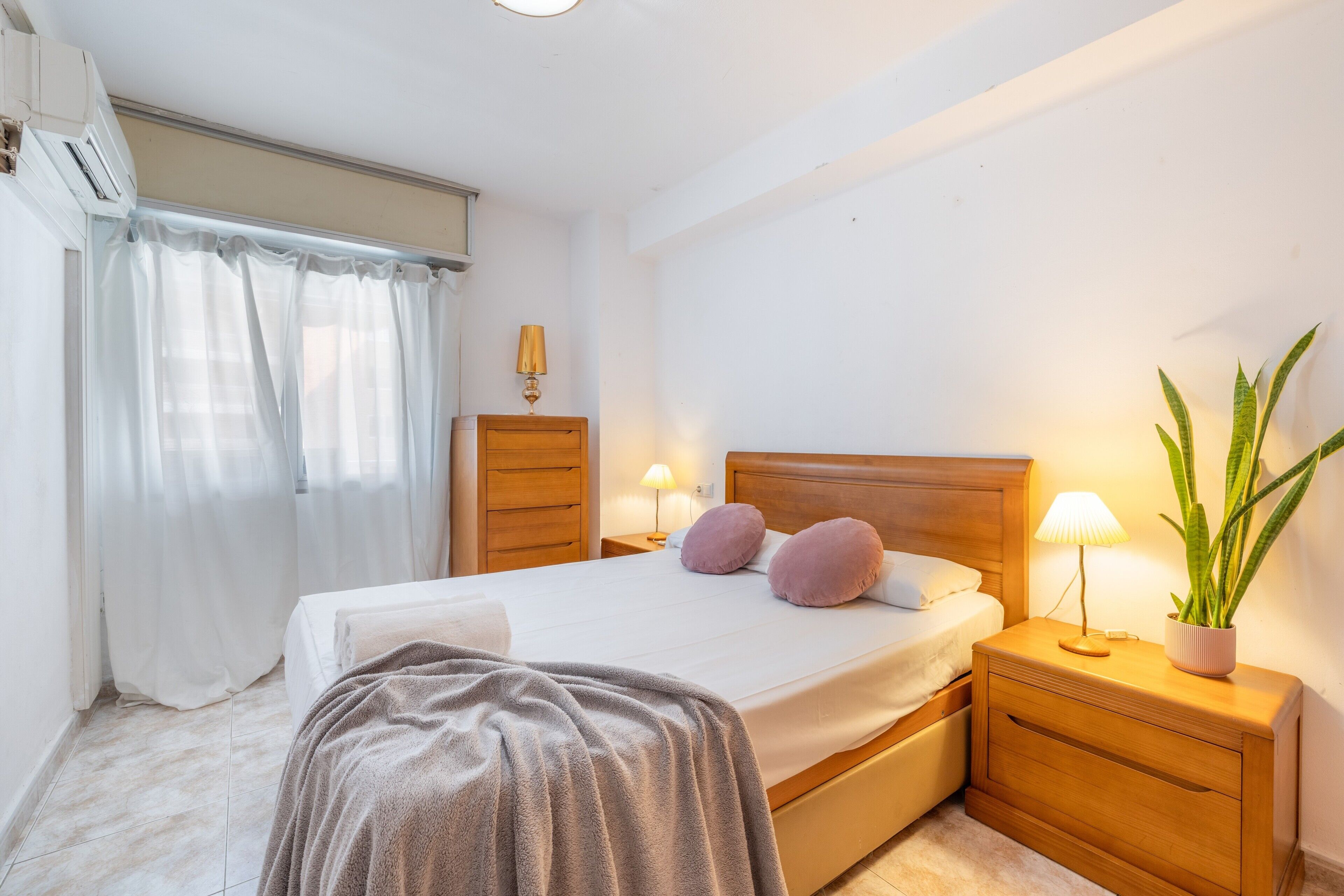 A bedroom with a bed, bedside tables, a lamp, a potted plant, and a window with curtains.