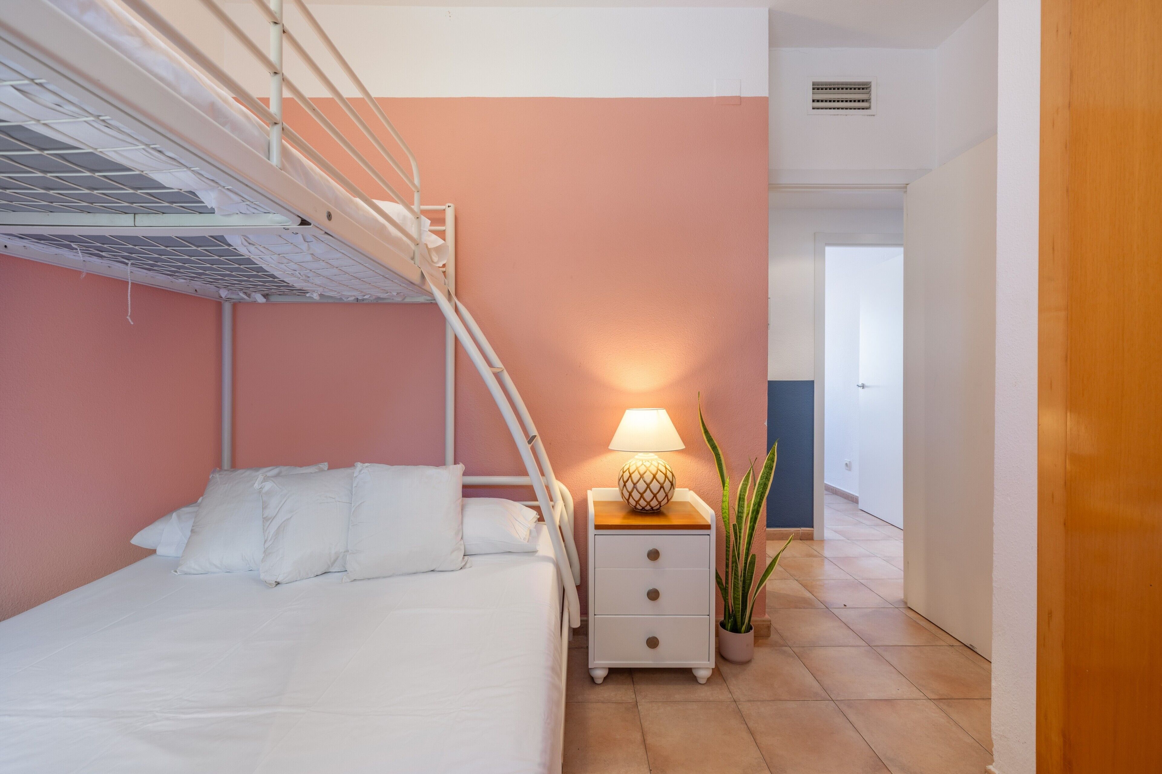 Een stapelbed met nachtkastje en lamp, een potplant en een gang naar een andere kamer.