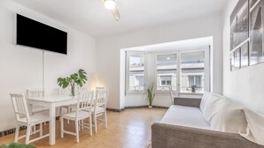 Ein modernes Wohnzimmer mit einem weißen Sofa, einem Esstisch mit Stühlen, einem flachbildfernseher an der Wand und einem großen Fenster mit Blick auf Gebäude.