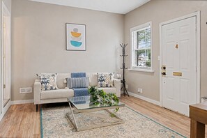 A living room with a sofa, a glass coffee table, a rug, a coat rack, and a door. - Walkerville 1BR Suite | Parking, Laundry & WiFi (Windsor)