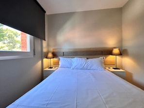 Una habitació d'hotel amb un llit, dues làmpades de nit, una finestra i un panell de fusta al capçal del llit.