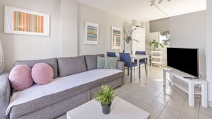 A modern living room with a grey sofa, a flat-screen TV, a dining area with a table and chairs, and a potted plant. - SunAndBed Penthouse Faro – Spectacular Sea Views, Pool, Sports Court (Salou)
