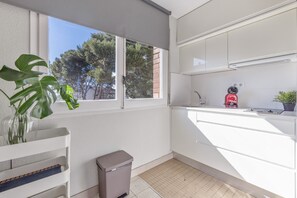 A modern kitchen with white cabinets, a window with a view of trees, a trash bin, and a potted plant. - SunAndBed Penthouse Faro – Spectacular Sea Views, Pool, Sports Court (Salou)