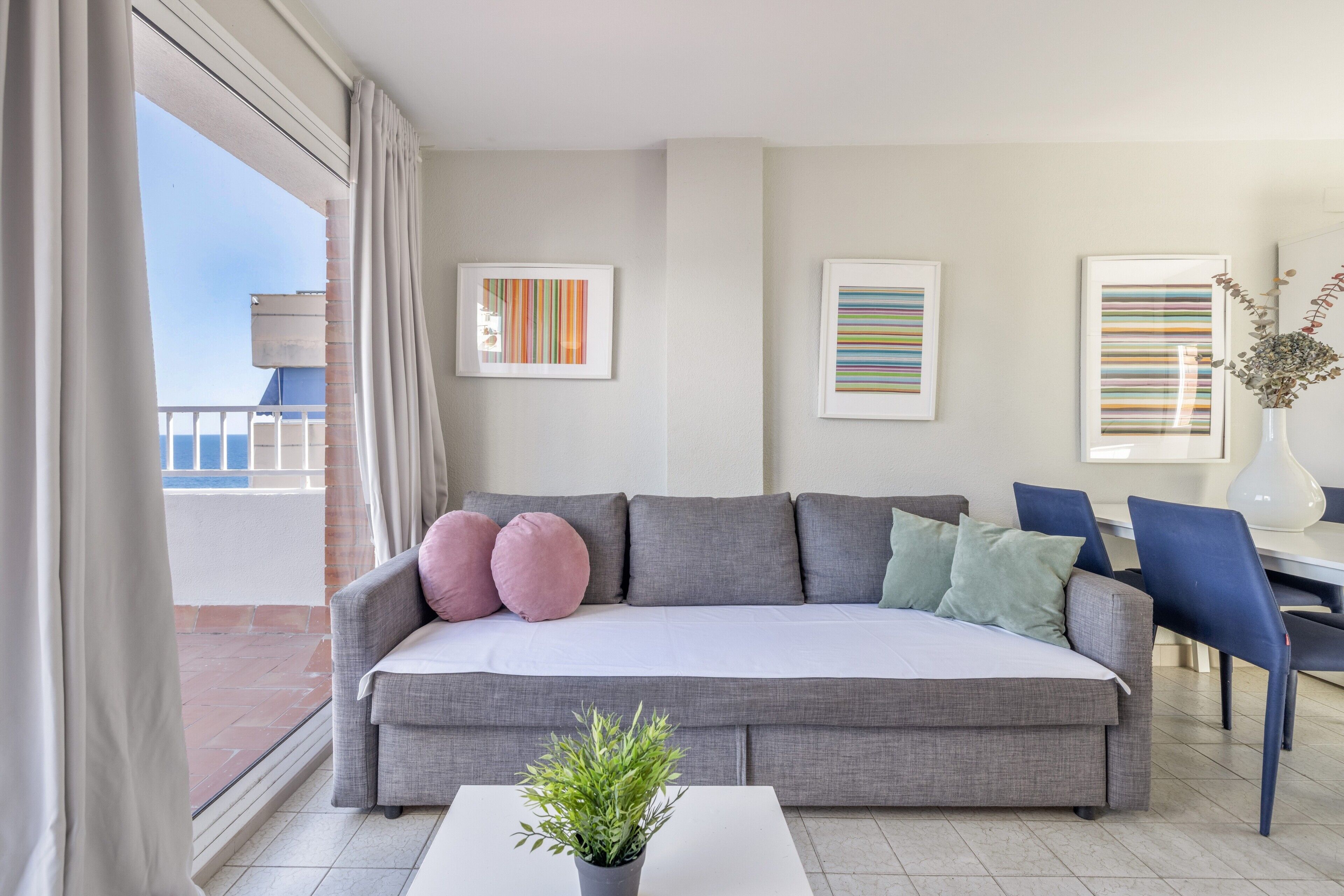 A modern living room with a grey sofa, a small white table, and a potted plant. There are framed artworks on the wall and a view of the outside.