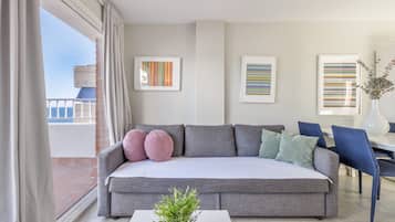 A modern living room with a grey sofa, a small white table, and a potted plant. There are framed artworks on the wall and a view of the outside.