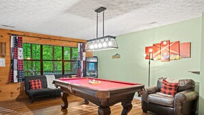Een pooltafel met een rode viltkleed, een leren bank met een ruitkussen, en een wandlamp.