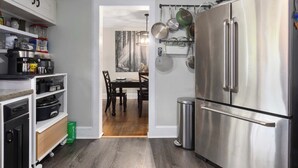 A kitchen with a stainless steel refrigerator, a wall-mounted pot rack, and a dining area with a table and chairs. - Spacious 4BR Home_Fenced Yard_Patio_BBQ Grill (Overland Park)