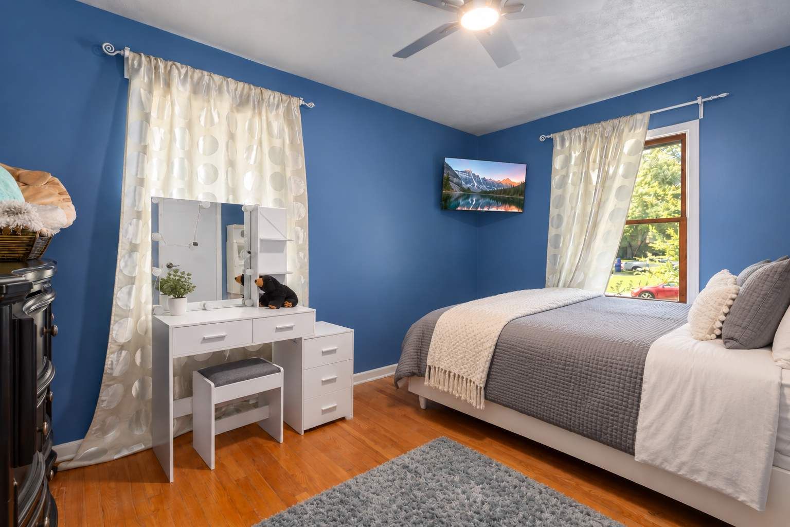 A bedroom with a bed, a desk with a mirror, a dresser, and a window with curtains.