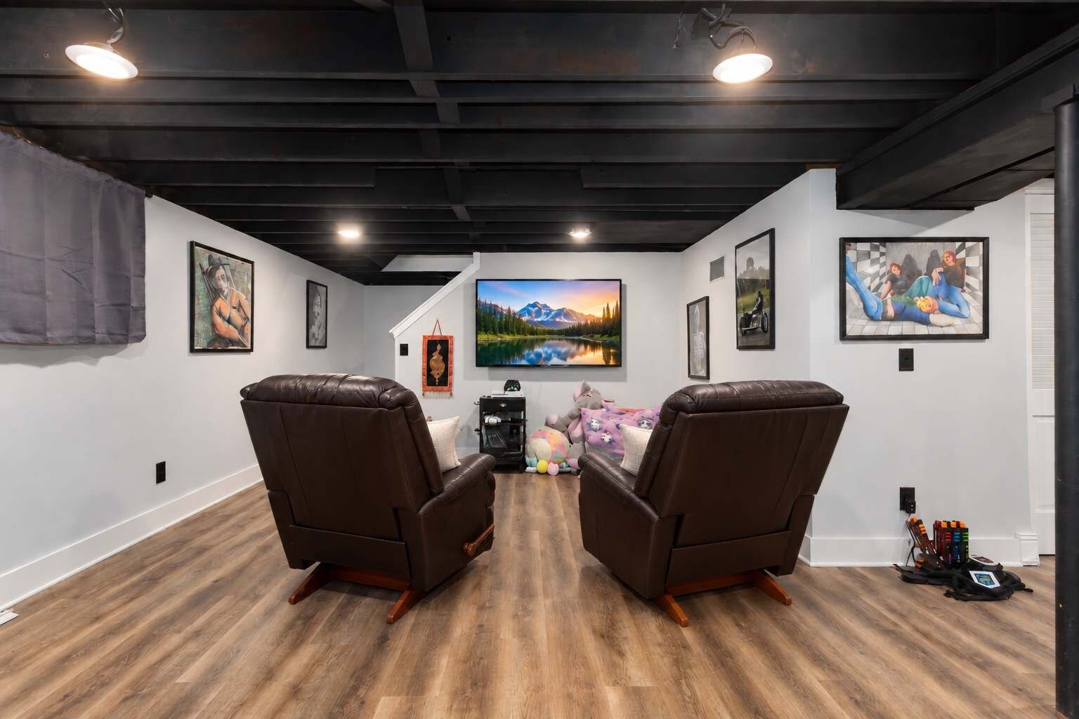 A room with two leather recliners facing a large flat-screen TV, wooden flooring, and artwork on the walls.