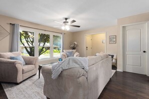 A living room with a beige sofa, a coffee table, a ceiling fan, and a large window with curtains.