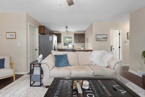 A living room with a beige sofa, a coffee table with text, a small side table, and a kitchen in the background.
