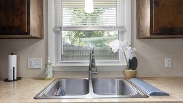 A kitchen with a double sink, wooden cabinets, a window with blinds, a hanging light fixture, a bottle of dish soap, and a potted orchid.