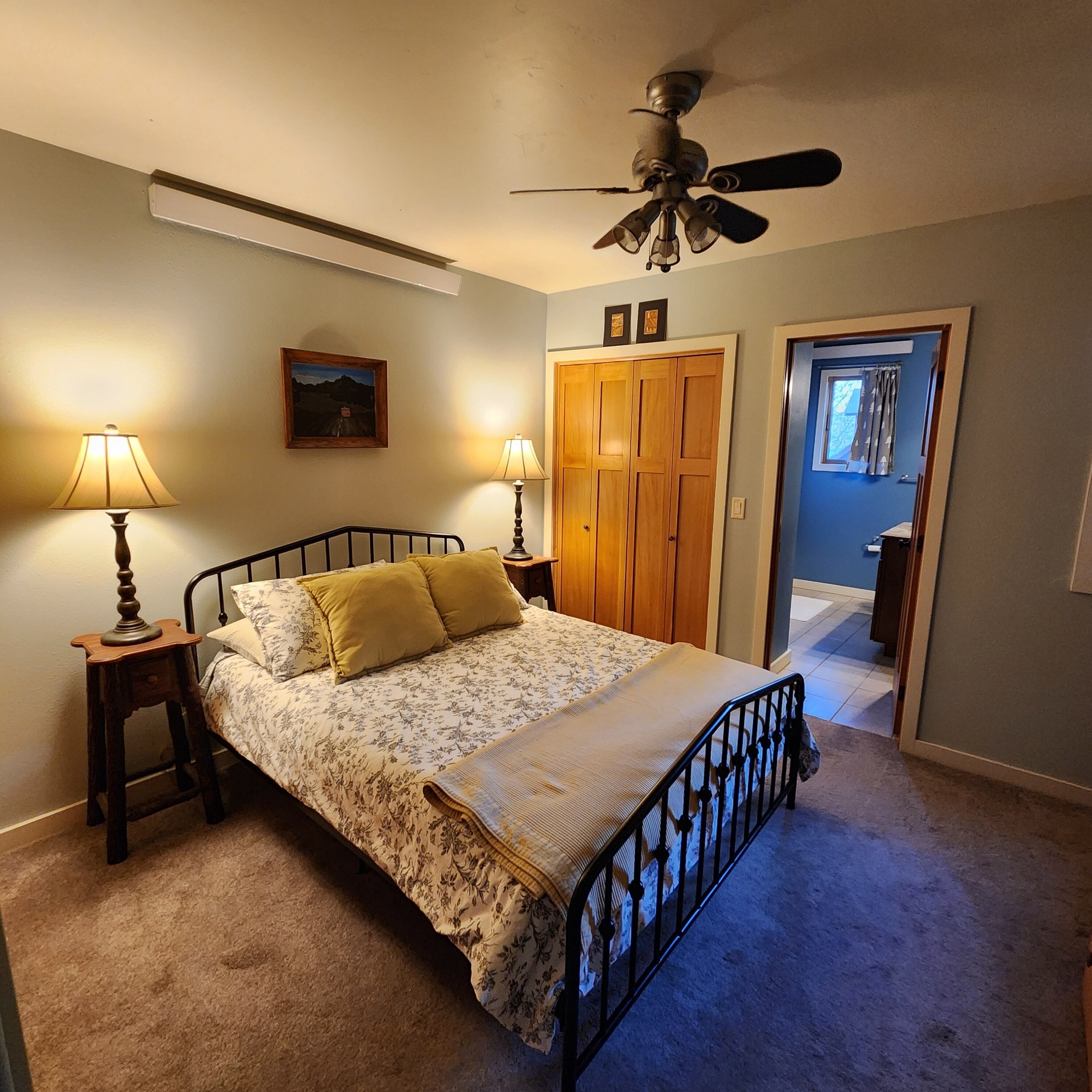 A bedroom with a bed, two bedside lamps, a ceiling fan, a door, and a painting on the wall.
