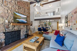 A modern living room with a stone fireplace, a television mounted on the wall, a dining area with a high ceiling, and a loft space. - Eagle Vail Retreat On Golf Course | Fire Pit (Avon)