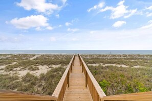 Ein hölzerner Steg führt zu einem Strand mit Dünen und vereinzelter Vegetation.