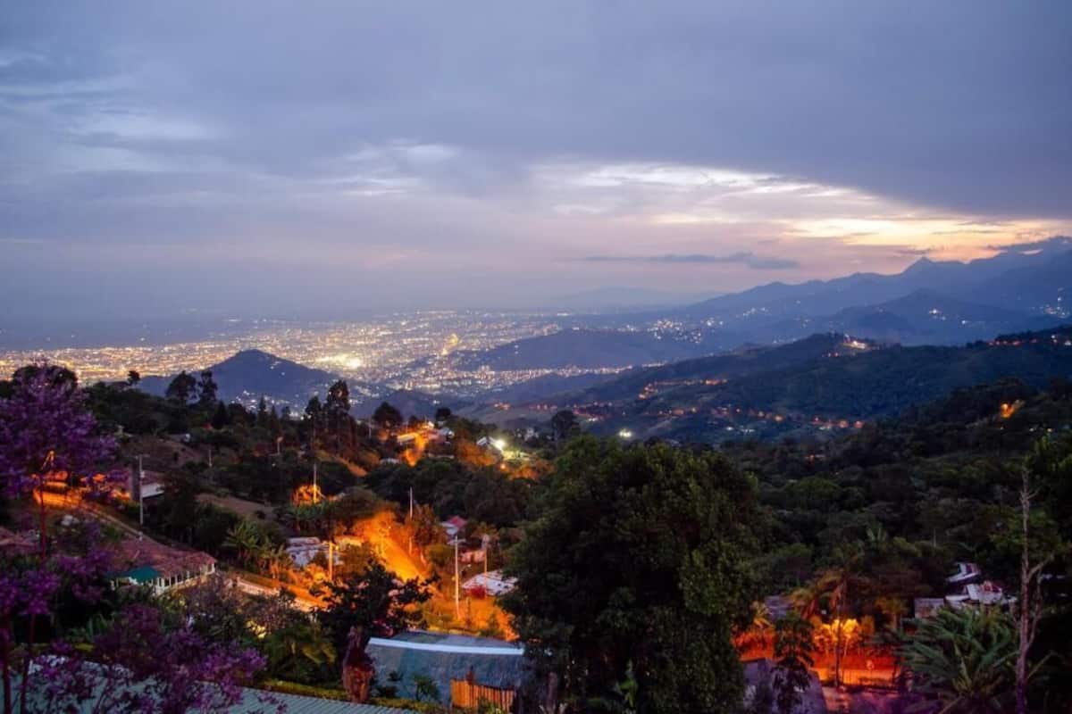 Vista panorámica de una ciudad al anochecer, con edificios iluminados y montañas a lo lejos.
