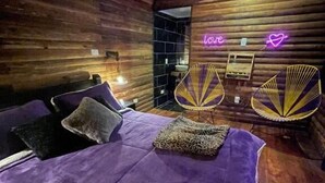 A room with wooden walls, a bed with a purple bedspread, two chairs with a fan, and a neon sign that says 'love'. - Romero Glamping y Cabañas (Cali)