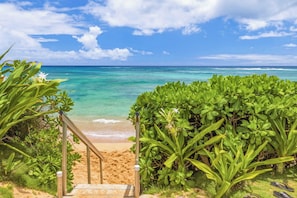 Ein Strand mit klarem türkisblauem Wasser, einem sandigen Ufer und einer Holztreppe, die zum Strand hinunterführt.