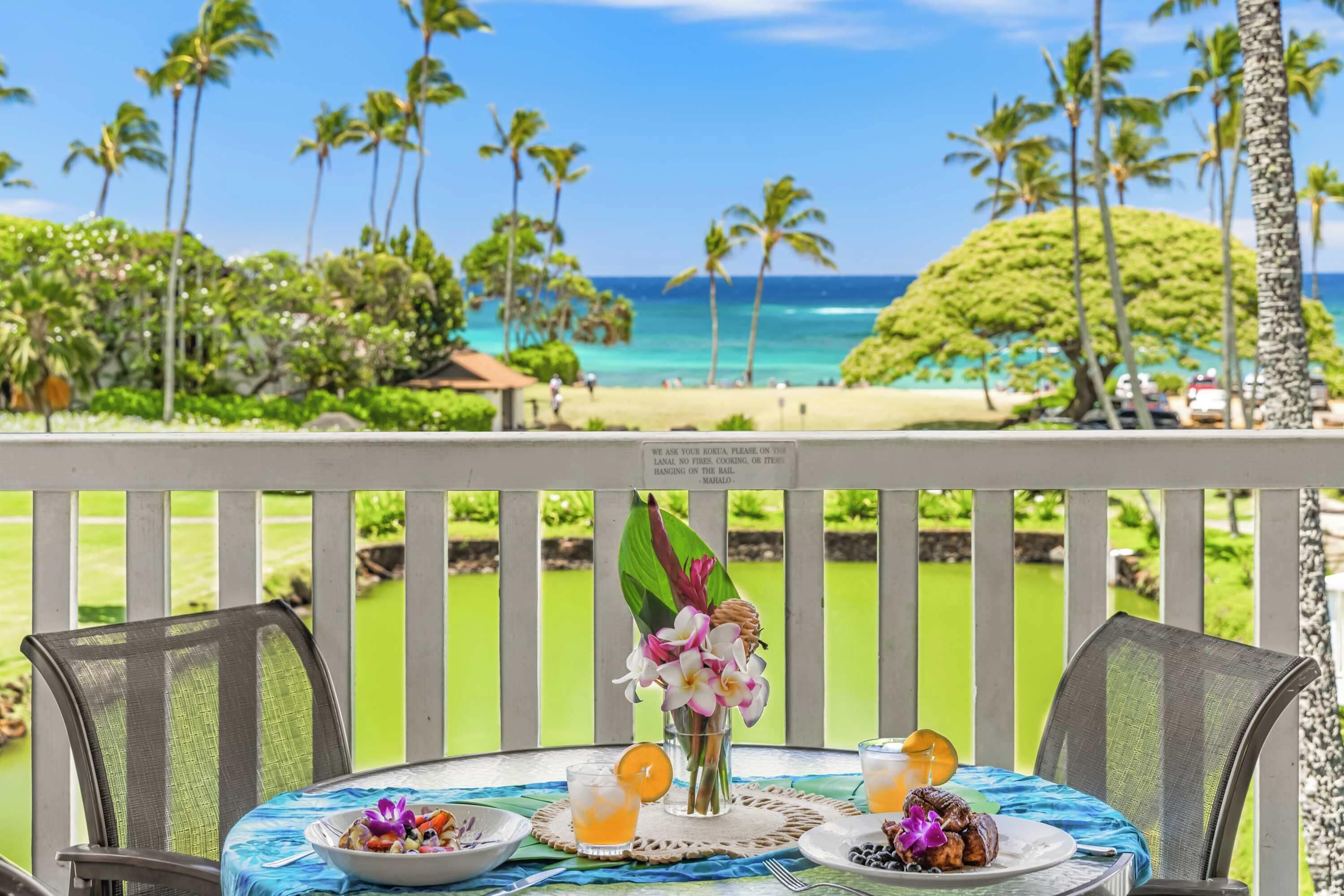 A table with a tropical drink, a bowl of food, and a vase of flowers on a balcony overlooking a beach with palm trees and the ocean.