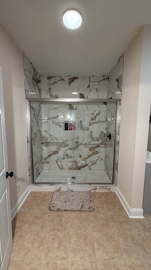 A bathroom with a glass shower enclosure, marble wall tiles, a glass door, and a beige floor.