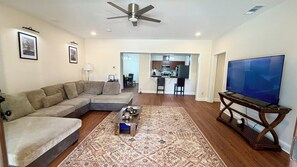 Ein Wohnzimmer mit Ecksofa, Couchtisch, Fernseher auf einem Lowboard, einem gemusterten Teppich und einem Deckenventilator.