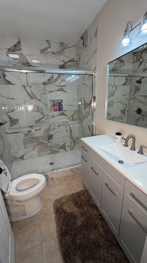 A bathroom with a glass shower enclosure, a toilet, a sink with a mirror, and marble-patterned walls.
