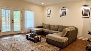 Ein Wohnzimmer mit Ecksofa, Couchtisch, gemusterten Teppichen und einer Tür mit Jalousien.