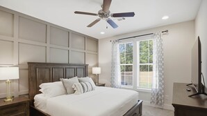 Quarto com cama, mesas de cabeceira, ventoinha no teto, janela com cortinas e televisão em um suporte.