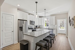 A modern kitchen with white cabinets, stainless steel appliances, a granite island with bar stools, pendant lighting, and a view of the outdoors. - Tiger Walk Townhouse (Auburn)