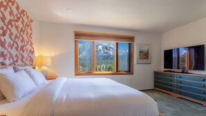 Ein Schlafzimmer mit einem Bett, einer Kommode, einem Fernseher, einem Fenster mit Bergblick und einer Lampe.