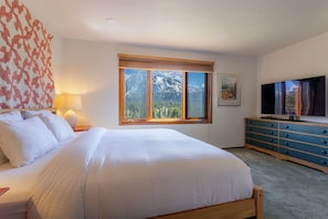 Ein Schlafzimmer mit einem Bett, einer Kommode, einem Fernseher, einem Fenster mit Bergblick und einer Lampe.