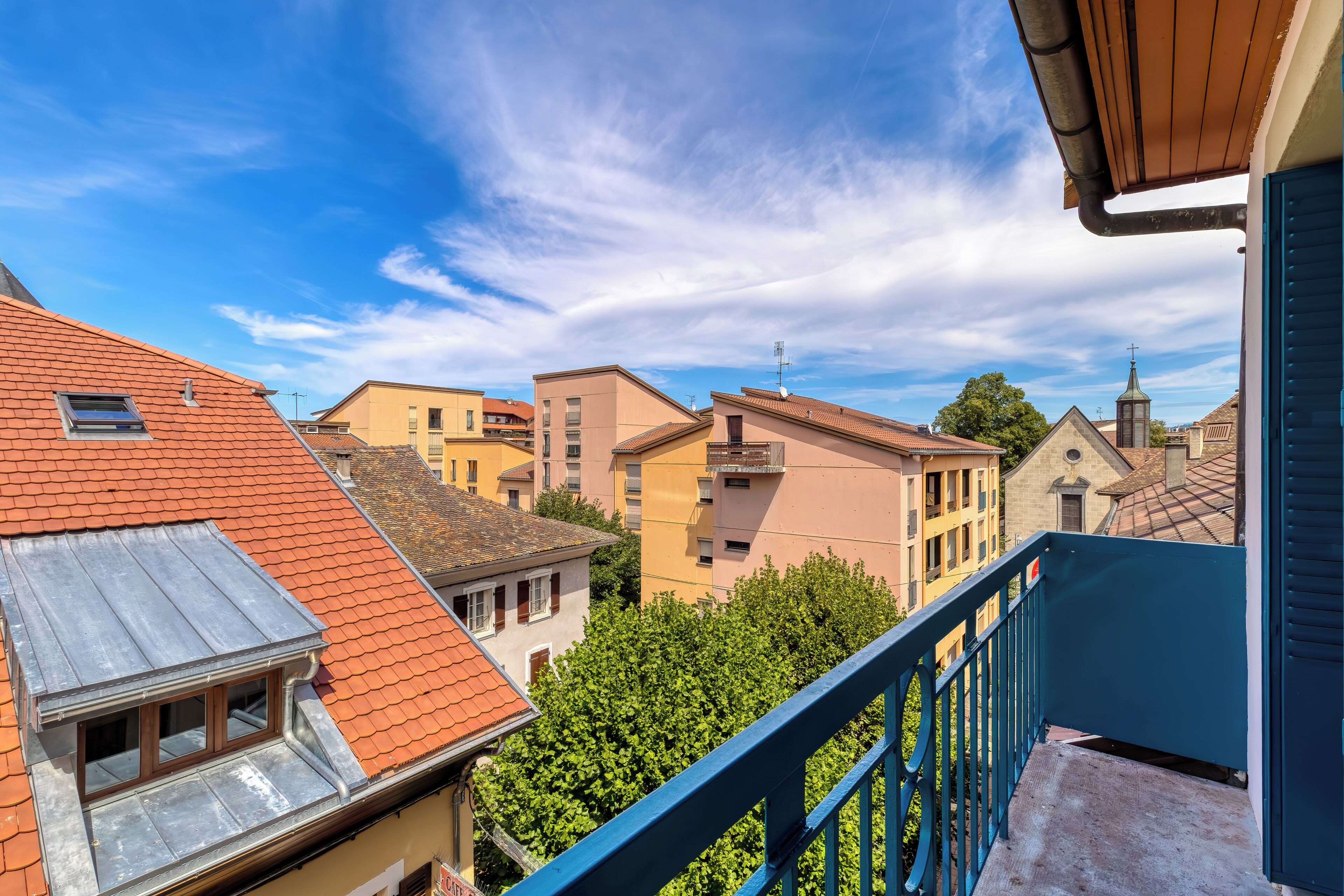 Ein Wohngebäude mit rotem Ziegeldach, blauer Balkonbrüstung und Blick auf benachbarte Häuser.