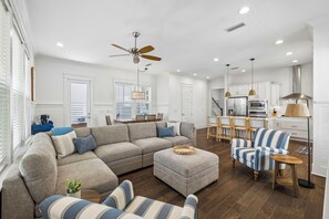A spacious living room with a sectional sofa, ottoman, and striped armchair, a dining area with a table and chairs, a kitchen with modern appliances, and a ceiling fan. - Newly Listed Home with Community Pool Access, LSV, Bikes, Grill and Firepit (Santa Rosa Beach)